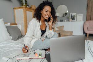 Woman sitting on her bed in front of her laptop making a phone call