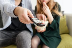 Two people sitting on a couch getting ready to change the channel while eating popcorn