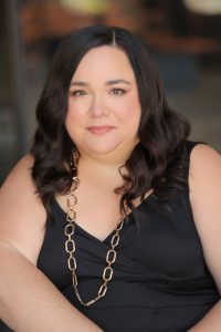 Human Voice Actor for AI Assistant with brown hair, a gold necklace, and a black v-neck shirt.