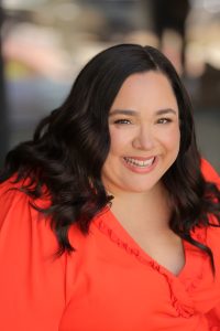 Professional award-winning Newscaster voice actor Maria Pendolino, wearing an orange blouse.